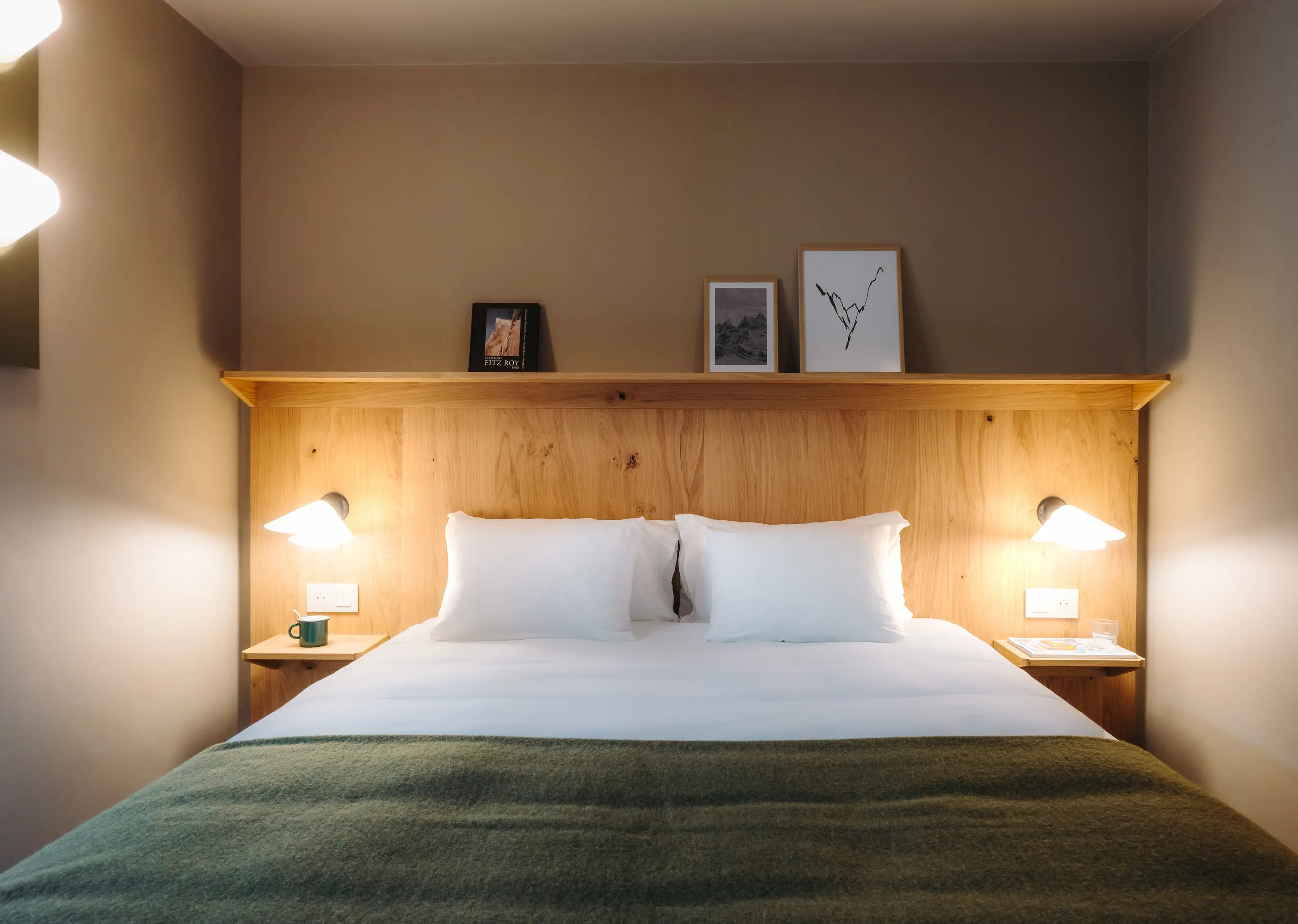 Chambre d'hôtel au style chalet contemporain avec un grand lit double en linge blanc et un plaid vert. La tête de lit en bois clair intègre deux liseuses et des tables de nuit. Une étagère au-dessus accueille des cadres avec des illustrations de montagnes et un livre "Fitz Roy". Ambiance chaleureuse et épurée.