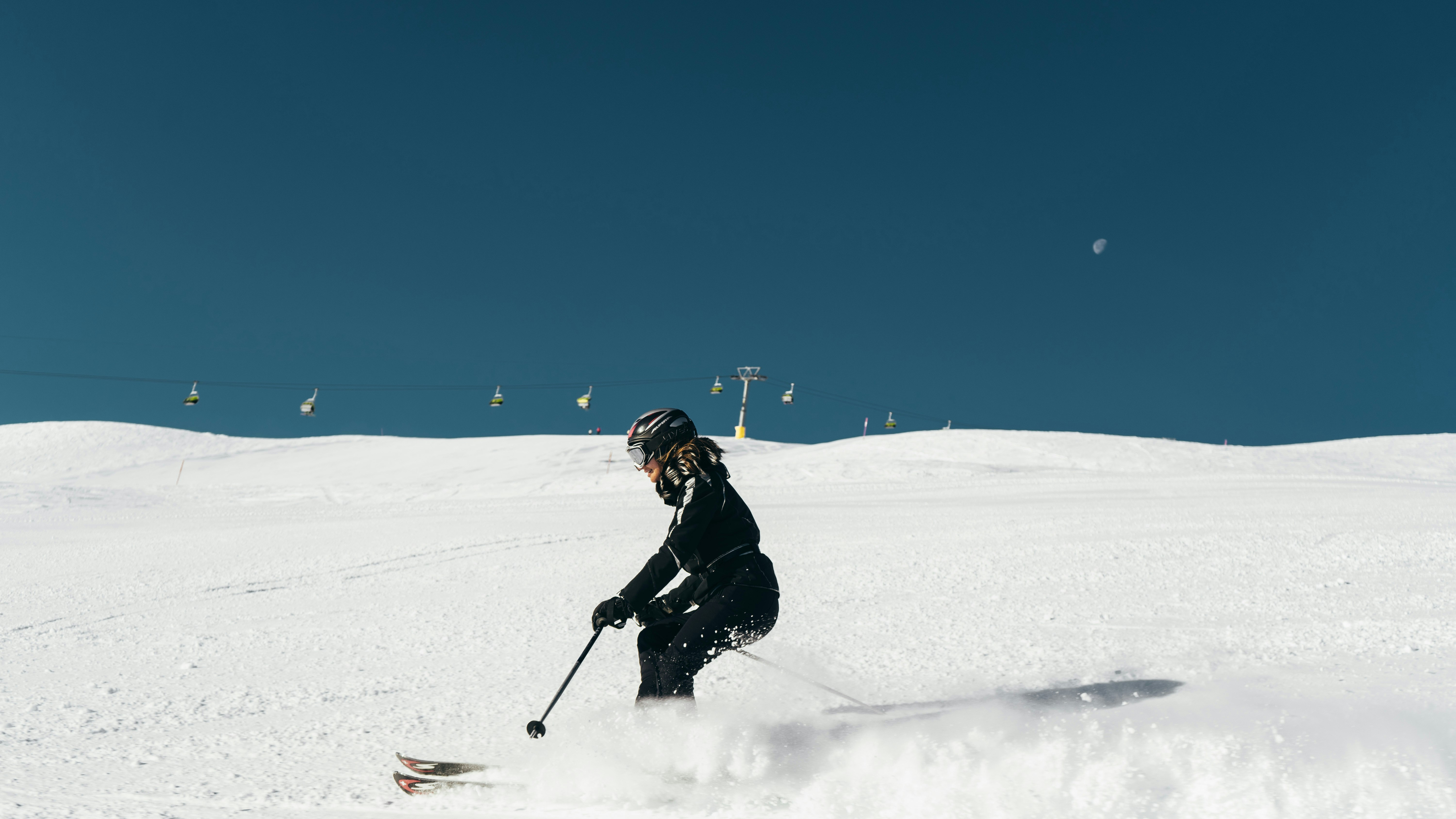 Skieuse en plein virage sur une piste damée avec télésiège et lune visible dans un ciel bleu