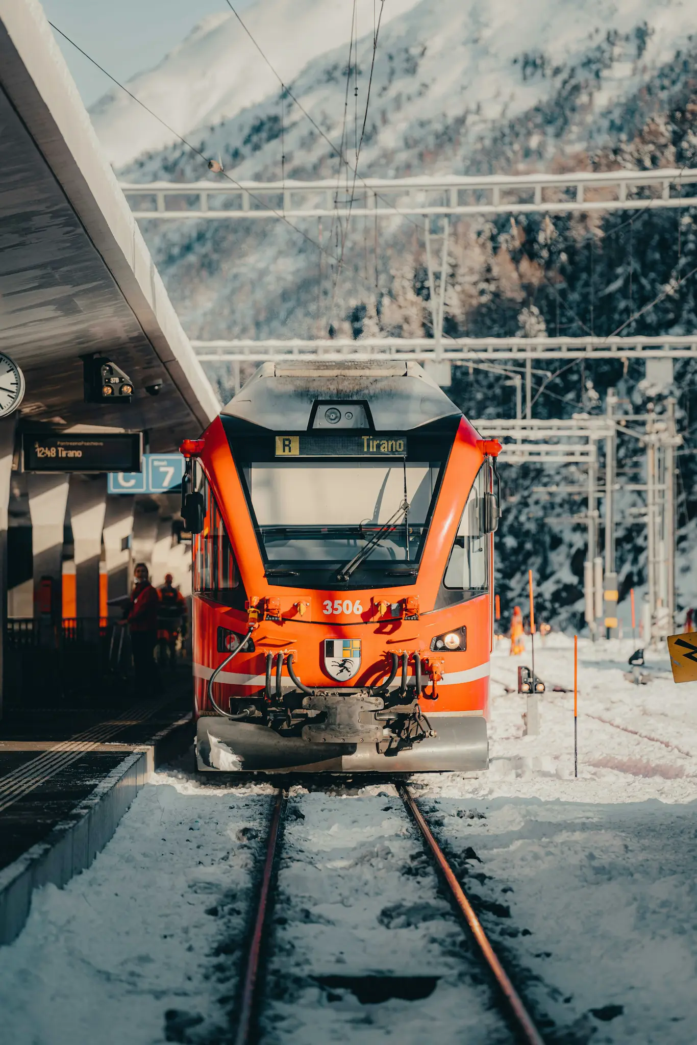 A red train stopped at the station, with snow-capped mountains in the background