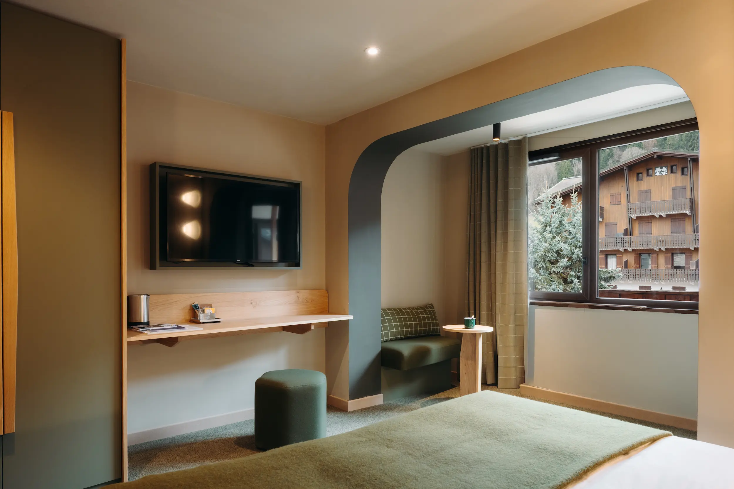 Chambre avec télévision murale, bureau en bois, pouf et alcôve donnant sur les chalets, Suite Garance